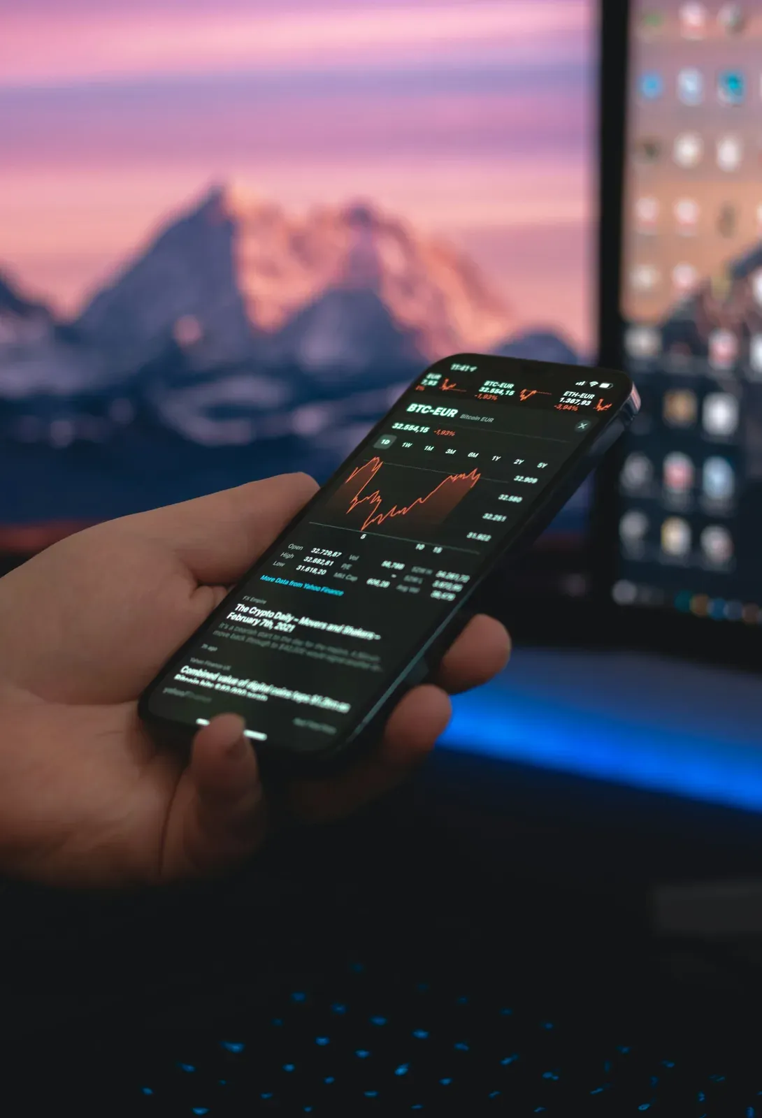 Hand holding a phone with a trading app in front of monitors