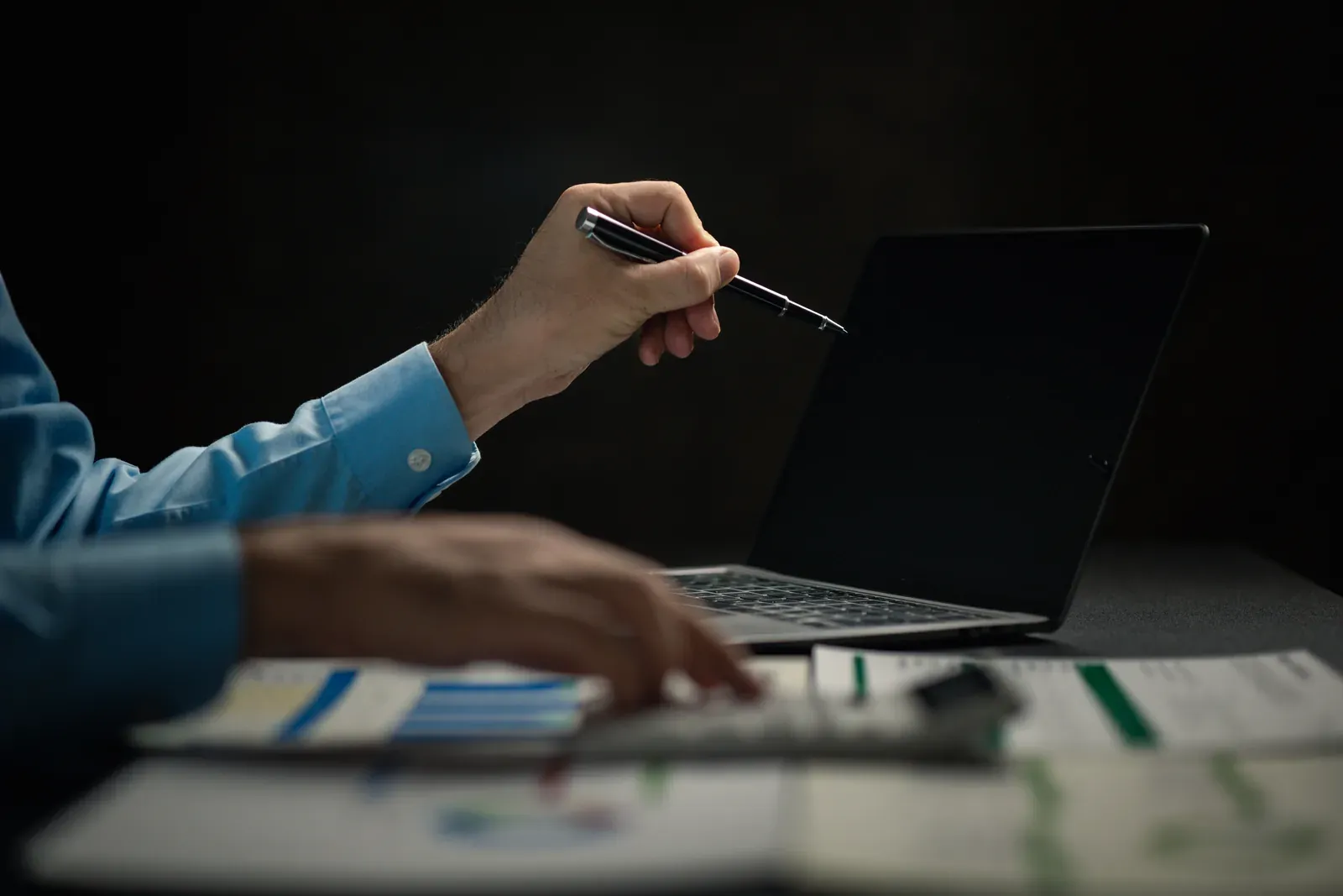 Analyst working at a laptop over printed charts in a dark room